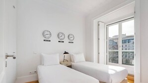 Habitación de hotel con dos camas, una ventana con vista a la ciudad y tres relojes en la pared que muestran diferentes zonas horarias.