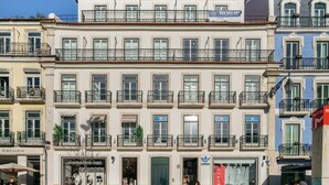 Un edificio de varios pisos con balcones, tiendas en la planta baja incluyendo una tienda de ropa de mujer y una tienda Adidas, y un número de teléfono visible en la fachada.