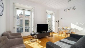 Una sala de estar moderna con un televisor de pantalla plana, sofás grises, una zona de comedor con una mesa y sillas blancas, y grandes ventanales con vistas a la ciudad.