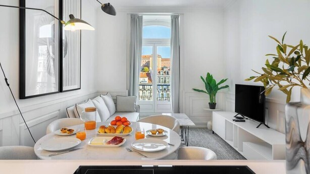 Ein modernes, helles Zimmer mit Esstisch, gedecktem Tisch für das Frühstück, einer Couch, einem Fernseher und einem Balkon mit Blick auf die Stadt.