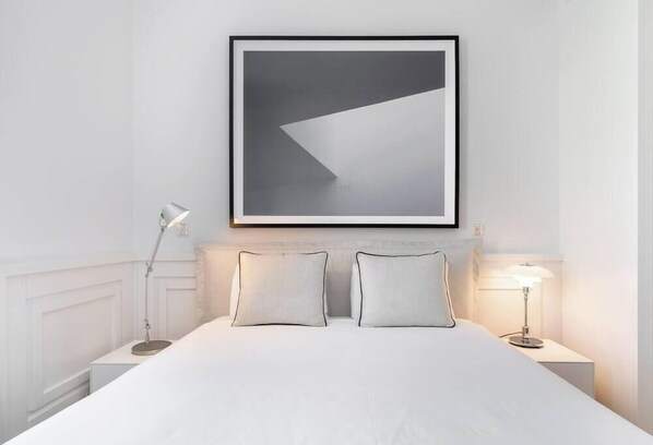 Una cama bien hecha con dos almohadas, una obra de arte abstracta enmarcada encima y dos lámparas de noche a cada lado.