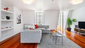 Ein modernes Wohnzimmer mit grauem Sofa, einem Couchtisch und einem TV-Schrank. Weiße Regale beherbergen dekorative Gegenstände, und ein großes Fenster sorgt für natürliches Licht.