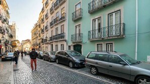 Eine Kopfsteinpflasterstraße gesäumt von mehrstöckigen Gebäuden, parkenden Autos und Fußgängern.