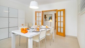 A modern dining area with a white table set for four, a hanging light fixture, and a view into a kitchen with wooden doors. - Family Holidays w/ S-Pool | AC | Elevator | Garage (Quarteira)