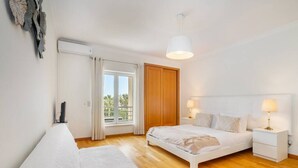A bedroom with a bed, bedside tables, a window with curtains, a wooden door, and a ceiling light. - Family Holidays w/ S-Pool | AC | Elevator | Garage (Quarteira)