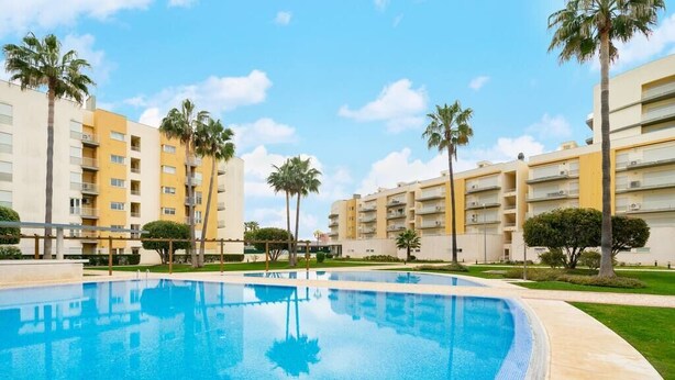 A swimming pool surrounded by apartment buildings with balconies, palm trees, and a clear sky. - Family Holidays w/ S-Pool | AC | Elevator | Garage (Quarteira)