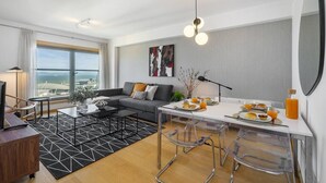 Ein modernes Wohnzimmer mit einer Couch, einem Couchtisch und einem Essbereich mit Stühlen. Ein großes Fenster bietet einen Ausblick, an der Wand hängt ein Gemälde und eine Lampe steht bereit.