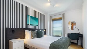 Ein modernes Hotelzimmer mit einem großen Bett, einem Nachttisch, einem Schreibtisch mit Stuhl, einem großen Fenster mit Aussicht und einem an der Wand montierten Fernseher.