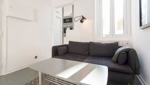 Uma sala de estar moderna, com um sofá cinzento, uma mesa de centro branca e uma área de cozinha visível através de uma porta aberta.