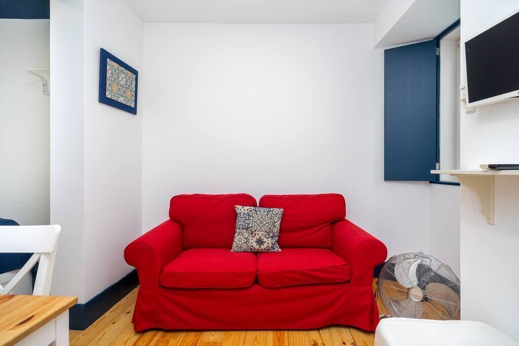 Een kleine, lichte kamer met een rode bank, één kussen, een ventilator en een houten vloer.