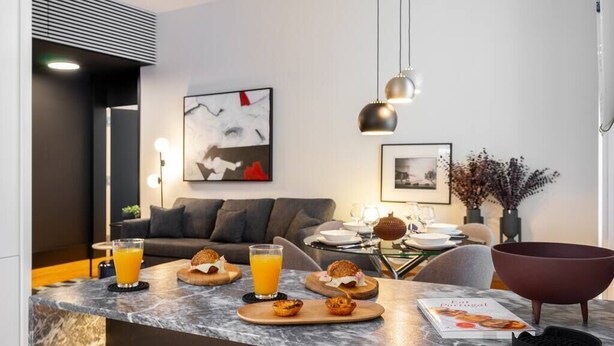 A modern kitchen with a marble countertop, a dining area with a table set for breakfast, and a living room with a sofa and artwork on the wall.
