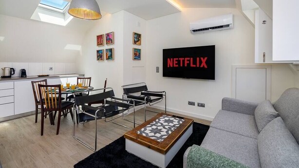 Ein modernes Wohnzimmer mit grauem Sofa, einem Couchtisch und einem an der Wand montierten Fernseher, der 'NETFLIX' zeigt.