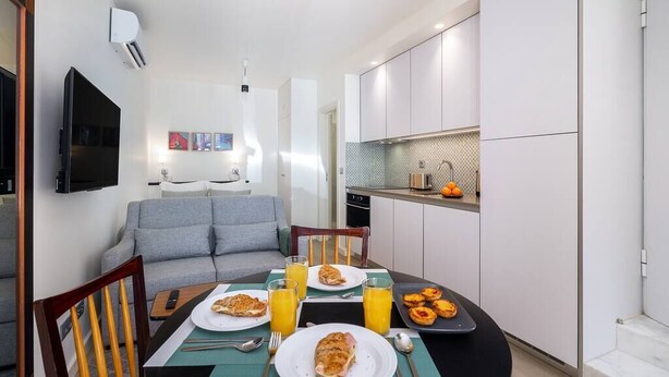 Een moderne keuken met witte kasten, een eettafel gedekt met eten en drinken, en een grijze bank op de achtergrond.