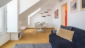 A modern living room with a dark sofa, a white dining table with chairs, and a staircase leading to an upper level. - Cozy Apartment in Lisbon’s Prime Area (Lisboa)