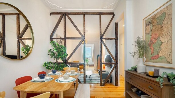 Uma área de refeições com uma mesa de madeira posta para quatro pessoas, rodeada por cadeiras e um banco. Há uma estante de madeira com objetos decorativos e um mapa na parede.