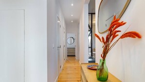 Um corredor com piso de madeira, um banco de madeira, um vaso verde com plantas secas e um espelho na parede.