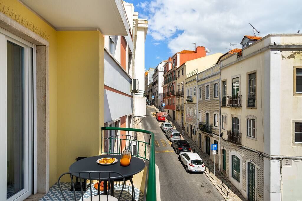 Una calle estrecha flanqueada por edificios, una mesa con sillas y comida en un balcón, y un coche estacionado.