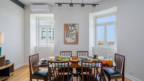 Une salle à manger avec une table ronde dressée pour six personnes, entourée de chaises en bois avec des coussins noirs. Un tableau est accroché au mur au-dessus de la table, et la pièce est dotée de grandes fenêtres sur un de ses côtés.
