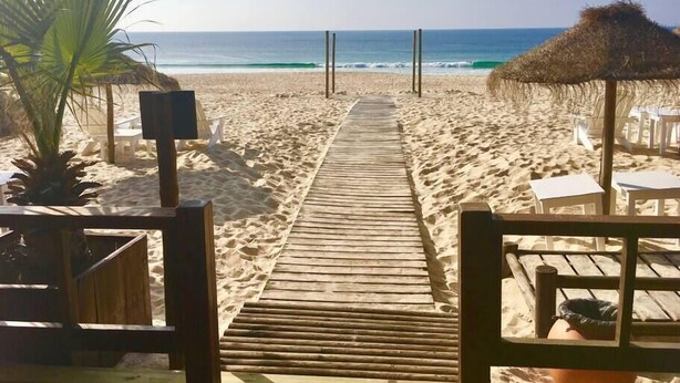 Een houten pad dat naar een strand leidt, met rieten parasols en witte stoelen.