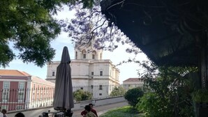 An outdoor cafe with tables and chairs, a building with a bell tower in the background, and a tree with purple flowers. - Family Apartment with Balcony in Graça (Lisboa)