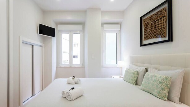 A hotel room with a large bed, two windows, a mounted TV, and a framed artwork on the wall. - Family Apartment with Balcony in Graça (Lisboa)