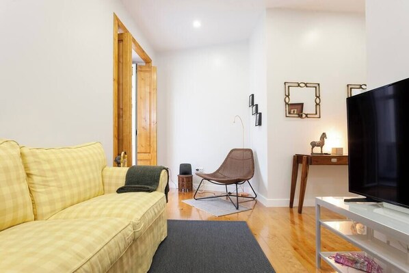 Un salon chaleureux avec un canapé jaune à carreaux, un fauteuil marron, un téléviseur posé sur un meuble blanc et une porte en bois donnant sur une autre pièce.