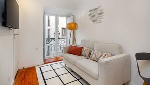 Un salon comprenant un canapé, un téléviseur, une lampe et donnant sur le balcon extérieur.
