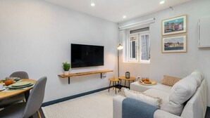 Een moderne woonkamer met een flatscreen televisie, een eettafel met stoelen, een bank, een klein bijzettafeltje en een raam met gordijnen.