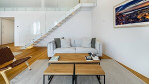 A modern living room with a glass staircase, a white sofa, a wooden coffee table, and a cityscape painting. - City View Penthouse Luxurious Living in Lisbon (Lisboa)