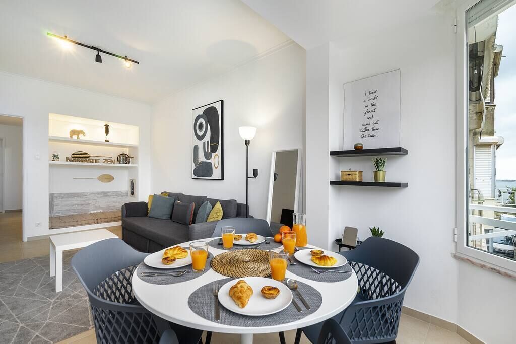 Uma sala de estar moderna com área de jantar posta para o café da manhã. Há dois sofás, uma mesa de centro e uma mesa de jantar com pratos, copos e um abajur.