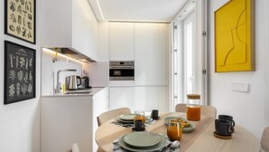 A modern kitchen with white cabinets, a built-in oven, and a coffee machine. There is a dining table with plates, cups, and a glass of orange juice.