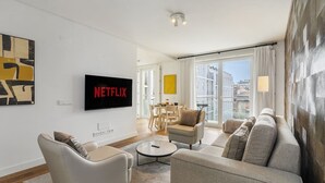 A modern living room with a flat-screen TV displaying 'NETFLIX', a sofa, a coffee table, and a view of the city through large windows.