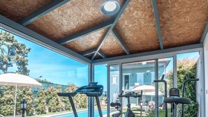 Uma academia em casa com esteira, elíptica e aparelhos de musculação, com grandes janelas e vista para o quintal.