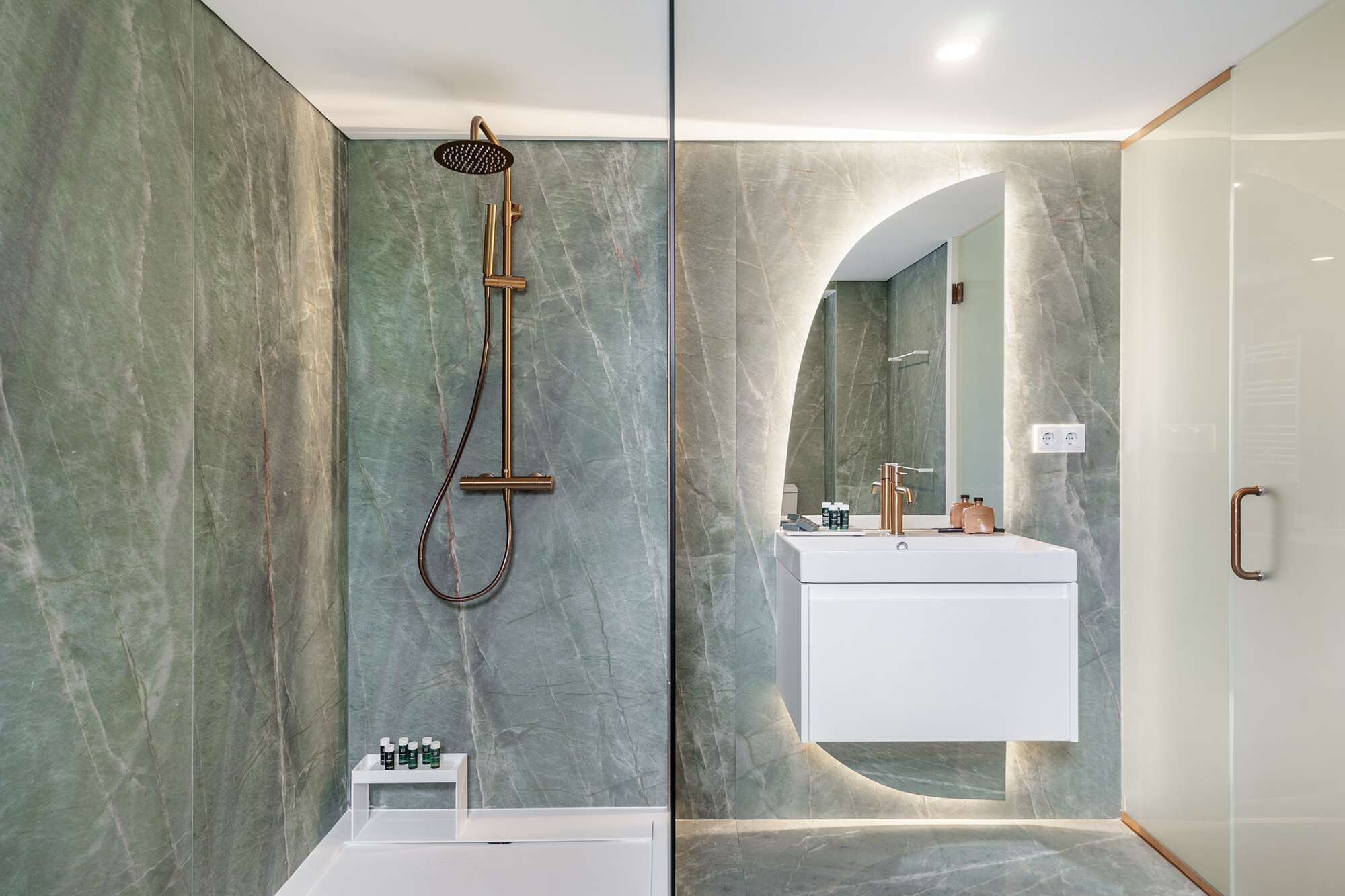 Ein modernes Badezimmer mit Marmorwand, freistehender Badewanne, Dusche mit Goldarmatur und Waschtisch mit Spiegel.