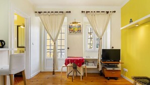 A room with wooden flooring, a dining table with a red tablecloth, a television on a wooden stand, and a window with curtains. - Central Alfama Hideaway perfect to explore Lisbon (Lisboa)