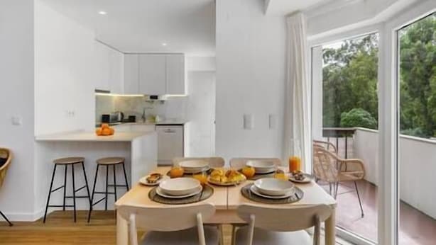 A modern dining area with a wooden table set for four, a kitchen in the background, and a large window with a view of greenery.