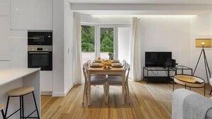 A modern kitchen with white cabinets and a dining area with a wooden table and chairs. There is a TV and a floor lamp in the living area.