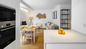 A modern kitchen with built-in appliances, a dining area with a wooden table and chairs, a sofa, and a small fruit display.