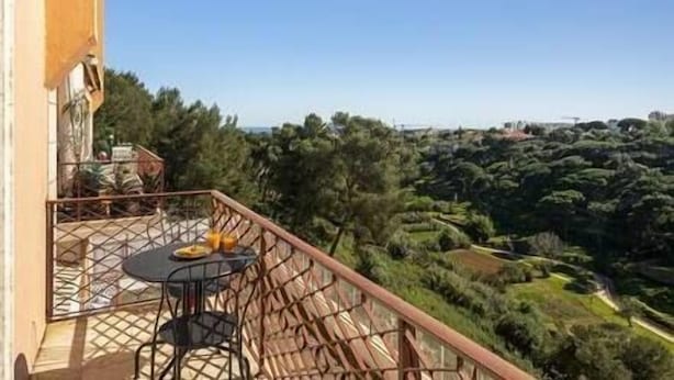 A balcony with a table and chairs, overlooking a lush landscape with trees and a distant view of a bridge.