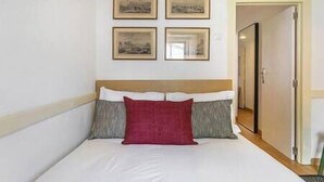 Cama bem arrumada com cabeceira de madeira, um travesseiro vermelho e dois cinzas. Quatro quadros emoldurados na parede acima da cama.