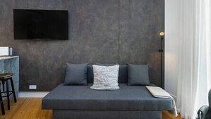 Ein modernes Schlafzimmer mit einer grauen Schlafcouch, einem Wand-Fernseher und einer Nachttischlampe.