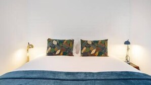 Ein Schlafzimmer mit weißer Wand, zwei Kissen mit Blättermotiv, einer blauen Decke und einer Nachttischlampe.