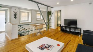 Ein modernes Wohnzimmer mit Holzboden, einem weißen Couchtisch mit Zeitschriften, einem Fernseher auf einem schwarzen Standfuß und einer Treppe mit Metallgeländer.