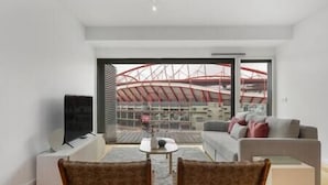 Un salón moderno con un sofá, dos sillones, un televisor y una mesa. Un ventanal ofrece vistas a un estadio de techos rojos.
