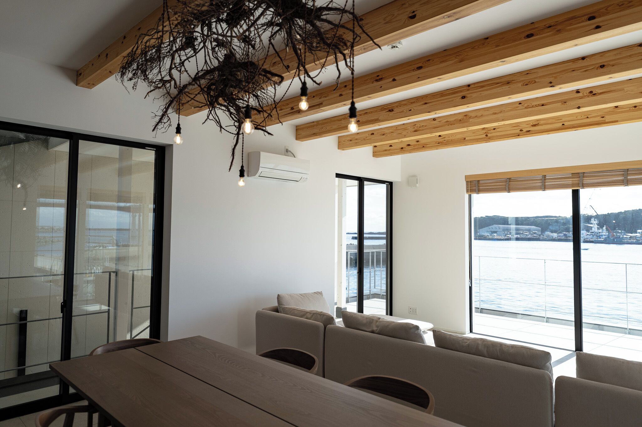 A modern living room with a wooden ceiling, large windows with a view of the sea, and a dining area with a wooden table and chairs.