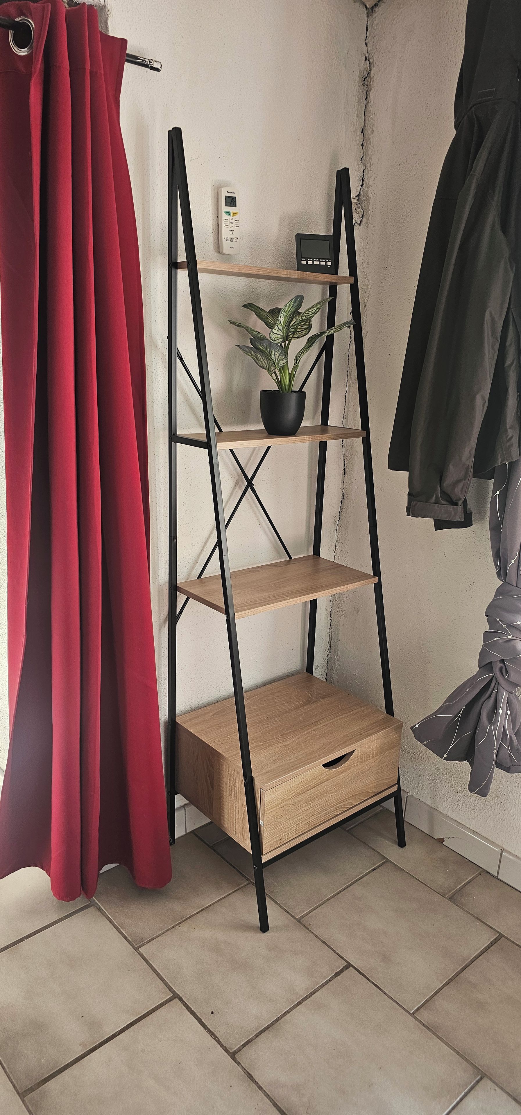 Een kamer met een zwarte metalen A-frame plankenwand, een potplant, een klein elektronisch apparaat en een rood gordijn.