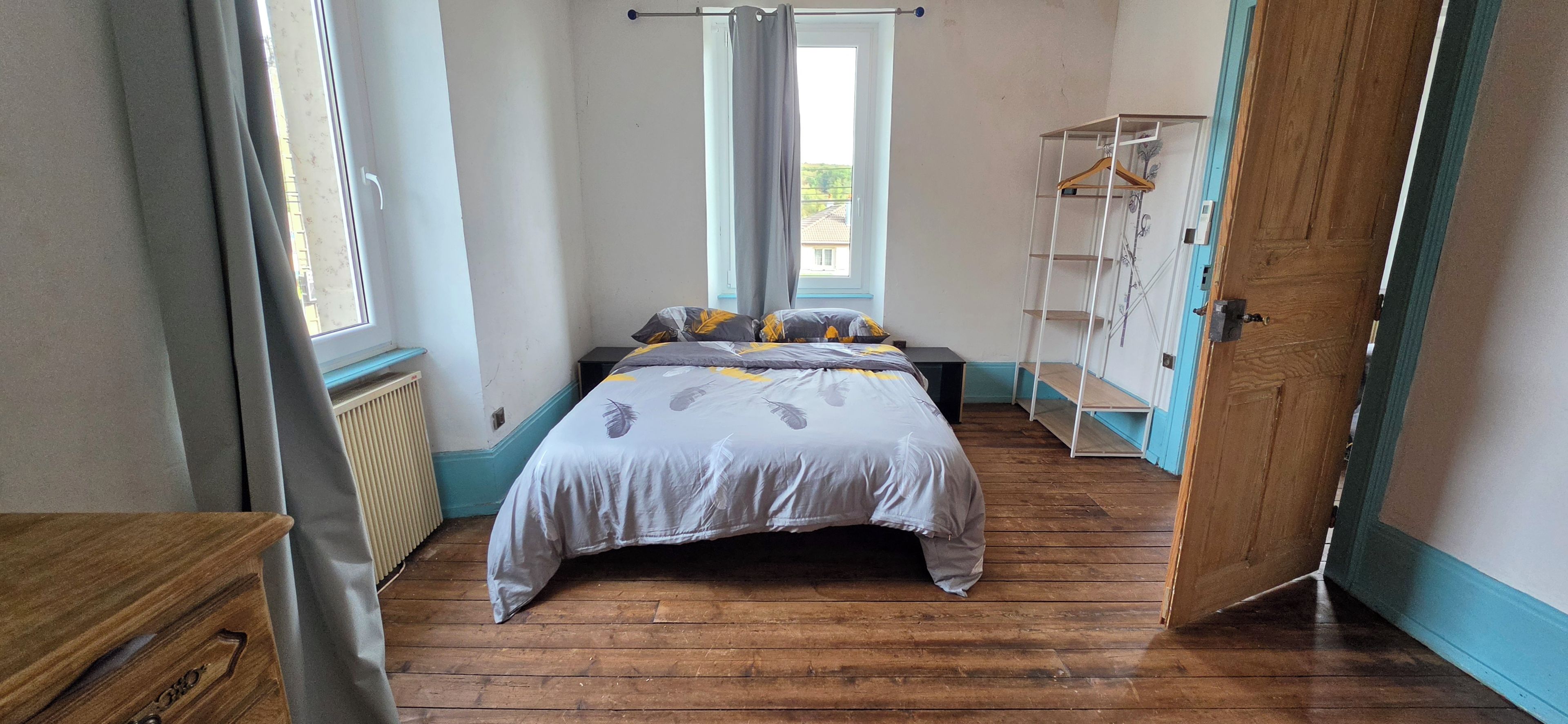 A bedroom with a wooden floor, a bed with a feather-patterned duvet, a radiator, a window with curtains, and a wooden wardrobe with a hanger.