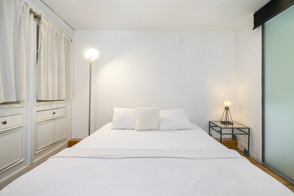 Un lit bien fait, avec une literie blanche, une lampe de chevet et une petite table sur laquelle se trouve une autre lampe.