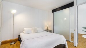 Un dormitorio moderno con una cama grande, una lámpara de pie, una mesita de noche con lámpara, y un ventanal corredero que da a un balcón.