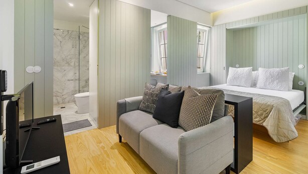 Un dormitorio moderno con un sofá gris, una cama con ropa de cama blanca, ducha de mármol y suelo de madera.
